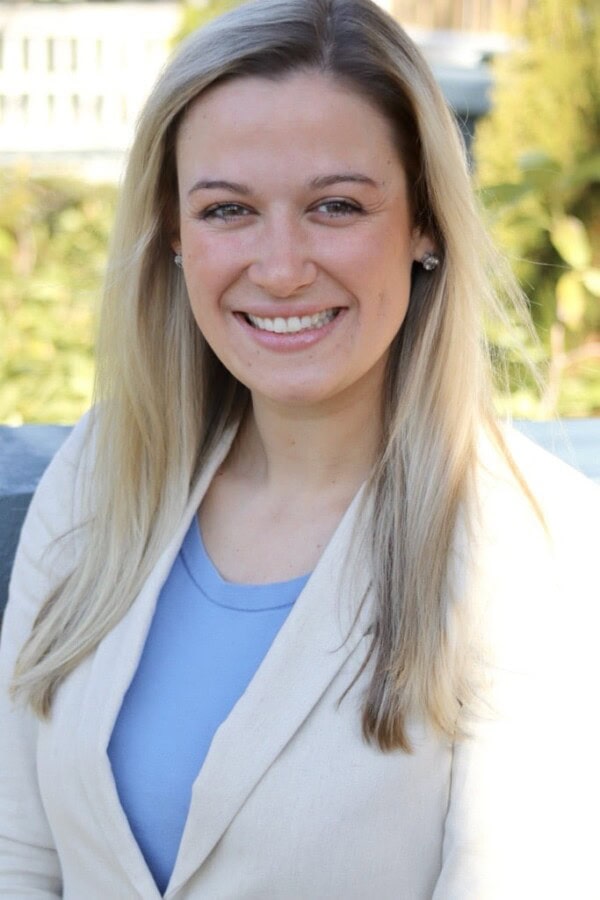 A woman with long blonde hair, wearing a light beige blazer over a light blue top, smiles while sitting outdoors with greenery in the background.