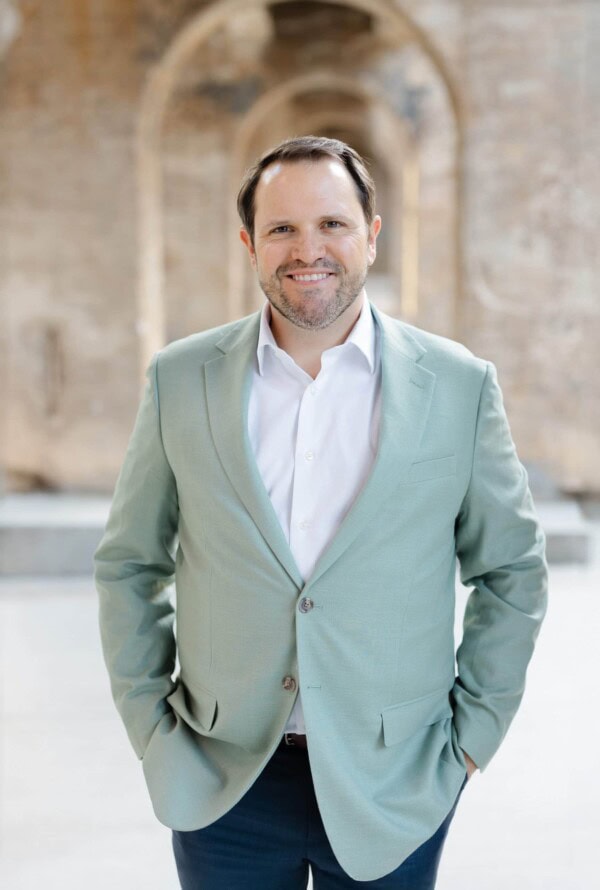 A man with short dark hair and a beard, wearing a light green blazer over a white shirt, stands smiling with his hands in his pockets in front of a blurred stone archway background.