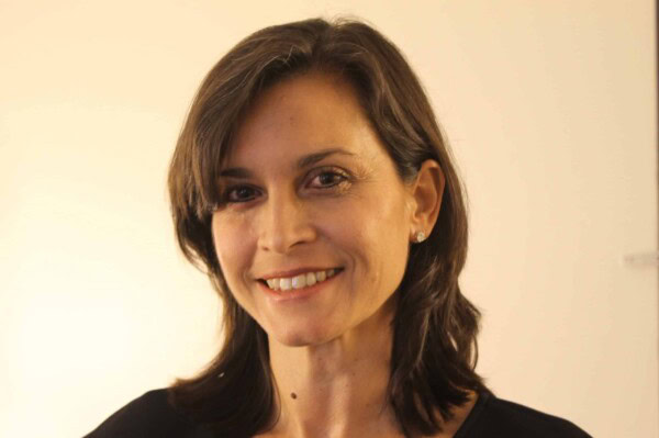A woman with shoulder-length brown hair and a side part smiles at the camera. She is wearing a black top and small stud earrings, standing in front of a plain, light-colored background.