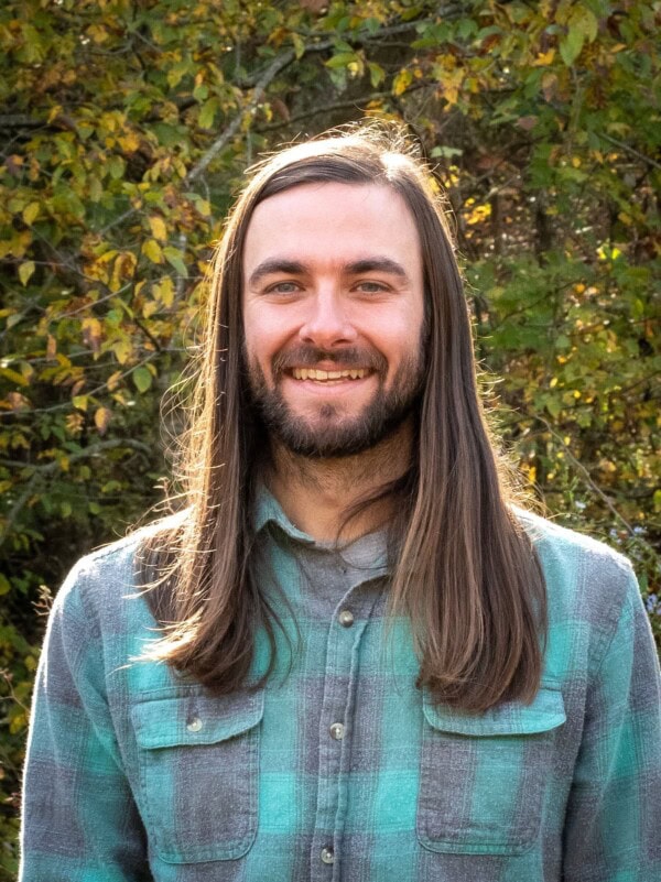 A man with long brown hair and a beard smiles outdoors in front of leafy trees, wearing a green and gray plaid shirt.