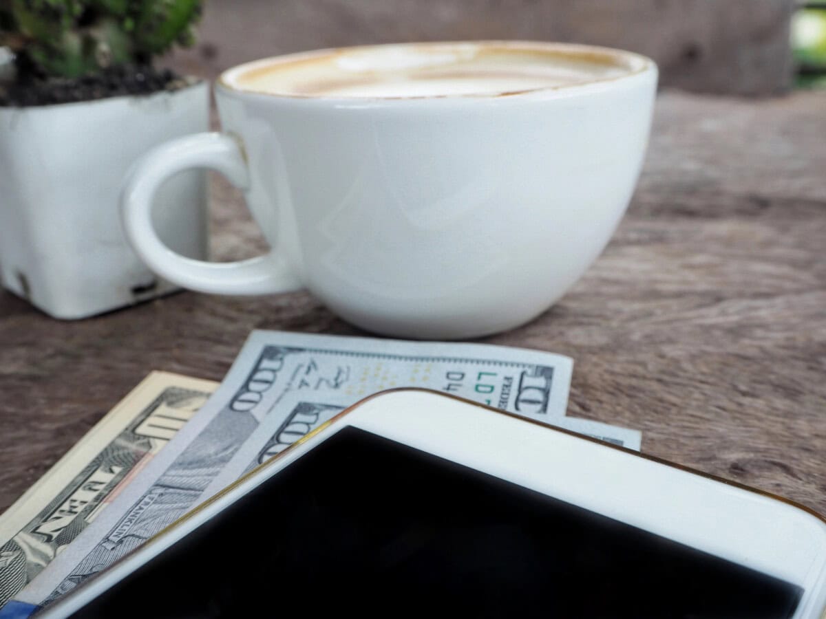A white coffee cup sits on a wooden surface next to a small potted plant, with several US dollar bills and a smartphone partially visible in the foreground.