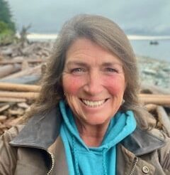A woman with long brown hair and a blue hoodie smiles at the camera. She is outdoors near a rocky shoreline with driftwood and water in the background under a cloudy sky.