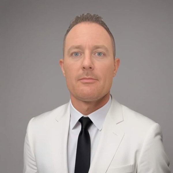 A man with short light brown hair wearing a white suit jacket, white shirt, and black tie stands against a plain gray background, looking directly at the camera with a neutral expression.