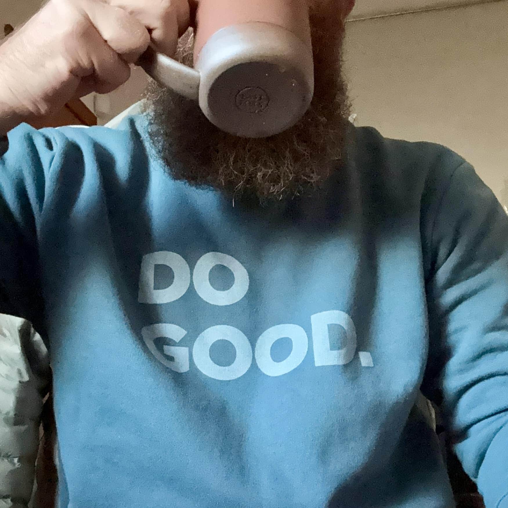 A person with a beard drinks from a mug, wearing a blue sweatshirt with the words DO GOOD. printed in large white letters.