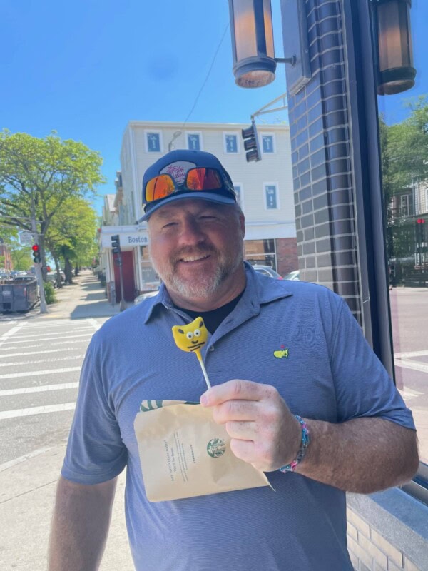 A smiling man in a blue shirt and cap stands on a sunny city street, holding a Starbucks bag and a yellow emoji cake pop.