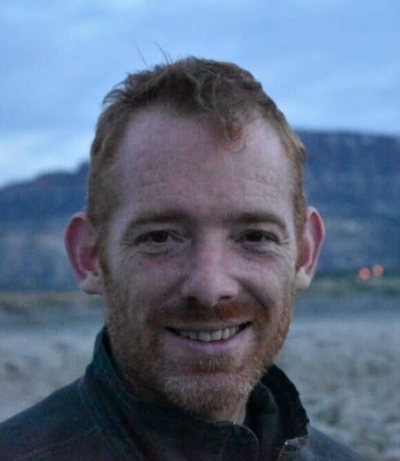 A man with short reddish hair and a beard smiles at the camera while standing outdoors, with a blurred mountainous landscape in the background under a cloudy sky.