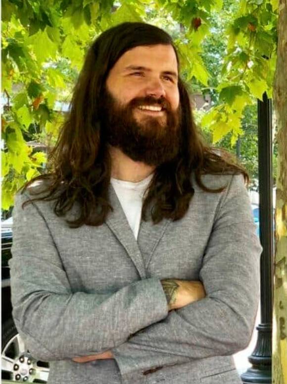 A man with long brown hair and a full beard smiles with his arms crossed, wearing a gray blazer over a white shirt. He stands outdoors in front of leafy green trees.