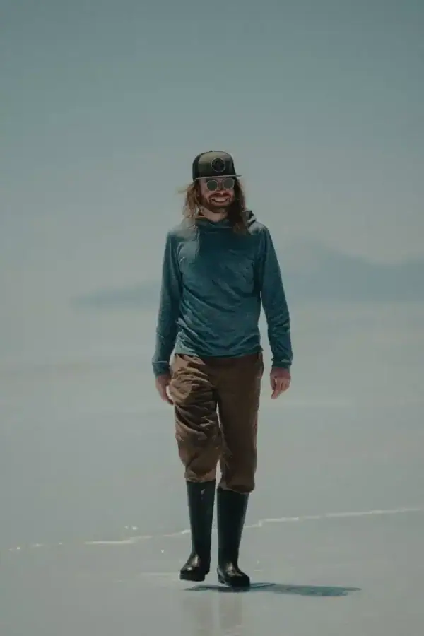 A person with long hair, wearing a cap, sunglasses, a blue long-sleeve shirt, brown pants, and tall black boots, stands on a reflective salt flat under a clear sky.