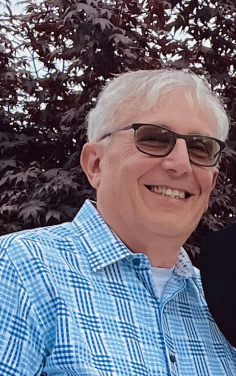A smiling older man with gray hair and sunglasses wears a blue and white checkered shirt, standing outdoors in front of leafy trees.