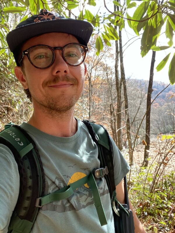 A person wearing glasses, a cap, and a backpack smiles while standing outdoors in a sunlit forest with trees and leaves in the background.