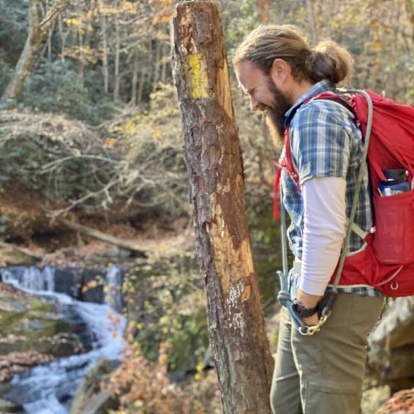 A man with a red backpack stands by a marked tree on a forest trail, smiling and looking down, with a small waterfall and autumn foliage in the background.