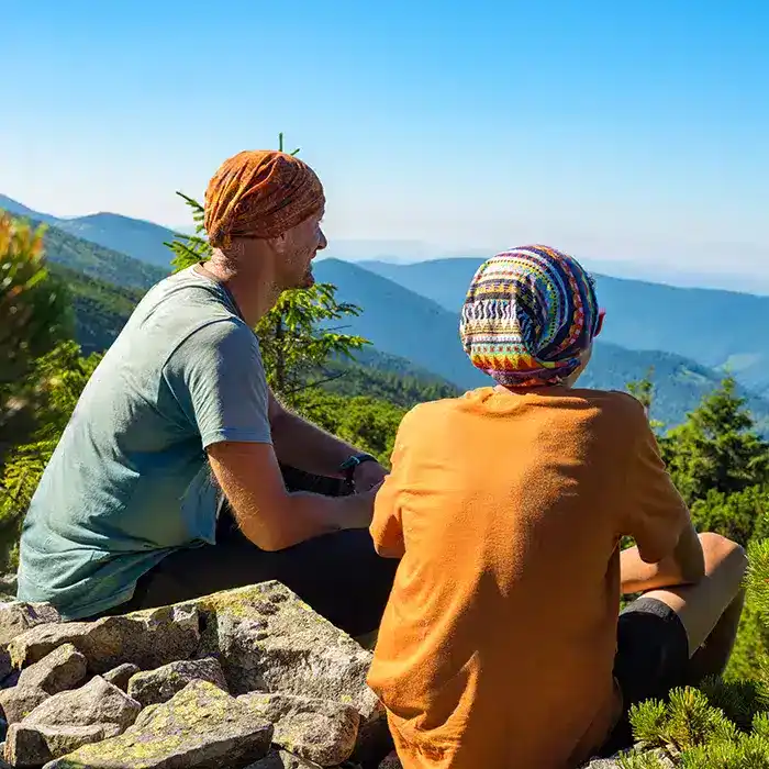 Two people wearing colorful headscarves sit on rocks, facing a scenic view of green hills and distant mountains under a clear blue sky.
