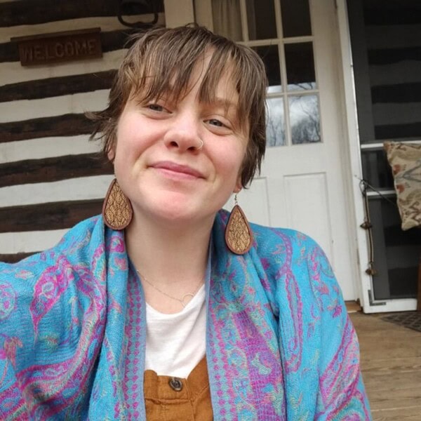 A person with short hair and large teardrop-shaped earrings smiles at the camera. They are wearing a blue patterned shawl. Behind them is a white door, a striped wall, and a “Welcome” sign.