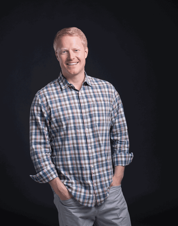 A man with short light hair smiles with his hands in his pockets. He is wearing a plaid long-sleeve shirt and light pants, standing against a plain dark background.