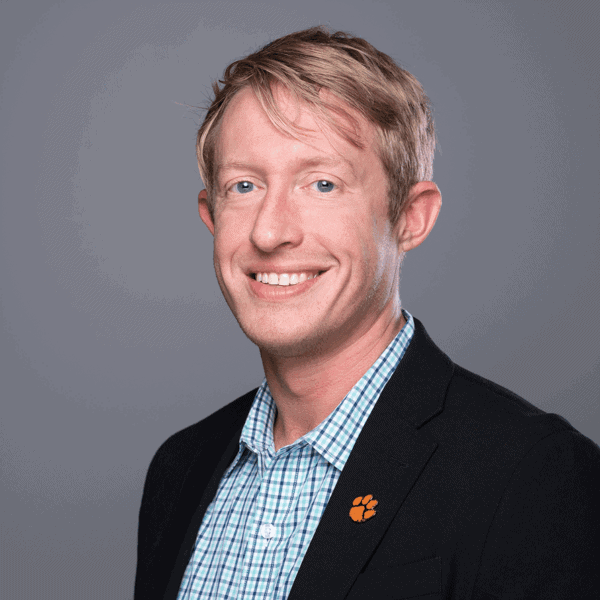 Smiling man with short blond hair wearing a black blazer, blue checkered shirt, and an orange paw pin on the lapel, posing in front of a plain gray background.
