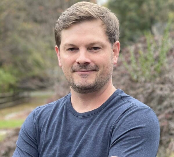 A man with short brown hair and a trimmed beard stands outdoors with his arms crossed, wearing a blue t-shirt. The background features trees and foliage, giving a natural setting.