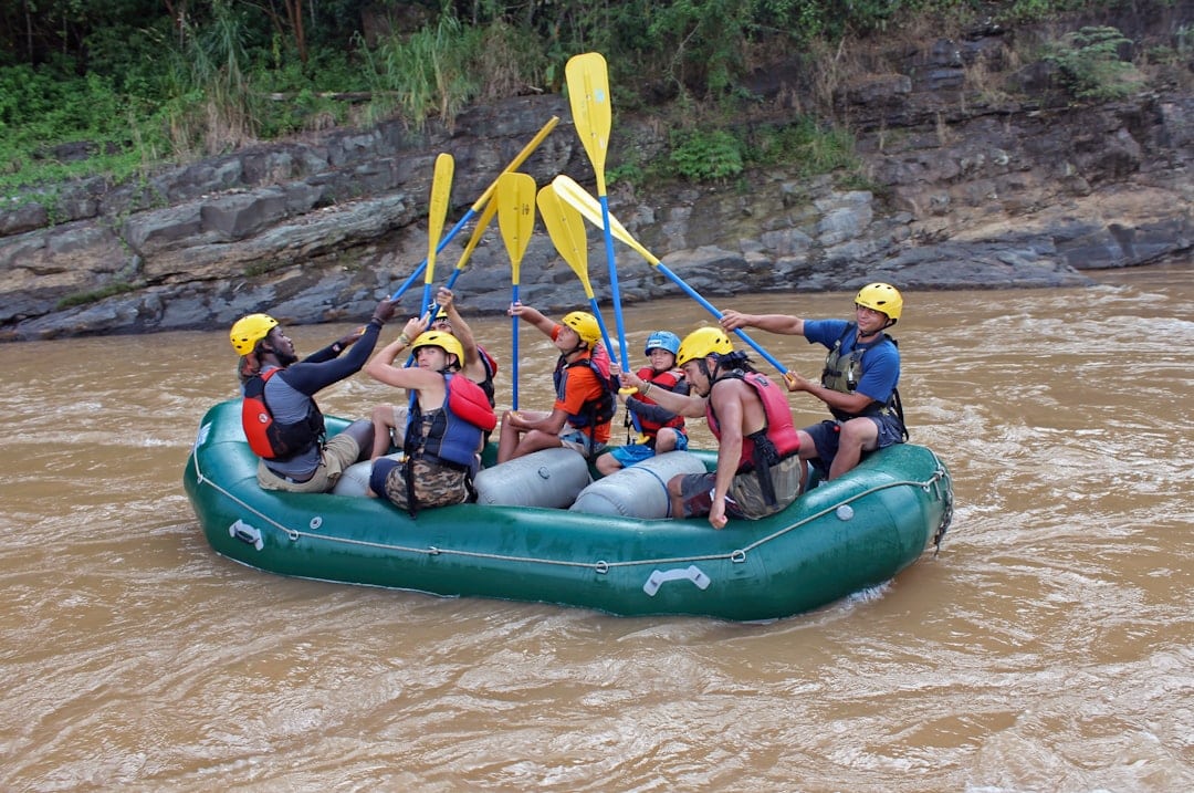 A group of people wearing helmets and life jackets raise their paddles together while rafting on a brown, fast-moving river in an inflatable green raft near rocky riverbanks.