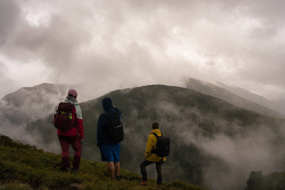 Three hikers wearing backpacks stand on a grassy hill, overlooking misty, cloud-covered mountains under a cloudy sky. The scene appears cool and foggy, with limited visibility of the distant peaks.