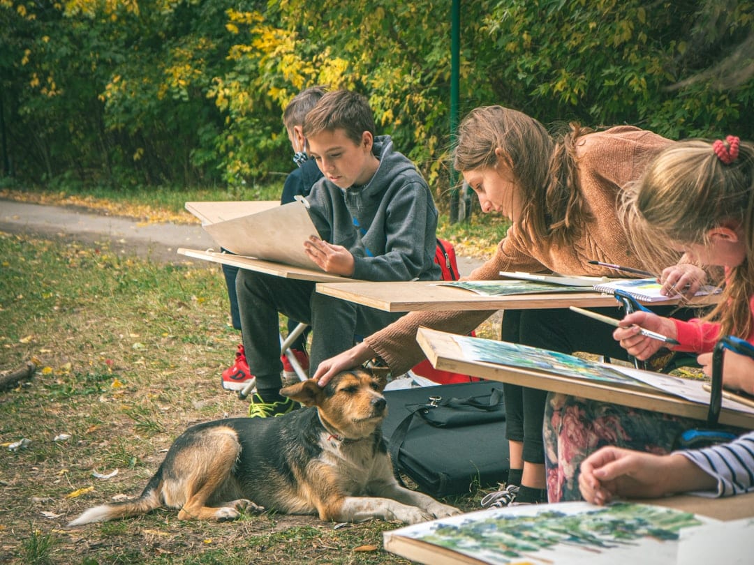 A group of children sit outdoors sketching on drawing boards. One girl pets a dog lying beside her, while others focus on their artwork. Trees with green and yellow leaves fill the background.
