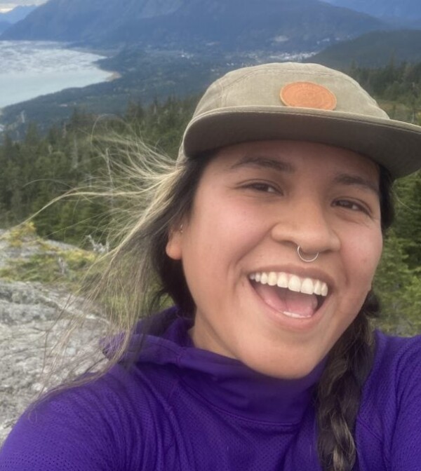A person with a nose ring, wearing a cap and purple jacket, smiles widely while taking a selfie on a mountain trail with a scenic view of forests, mountains, and water in the background.