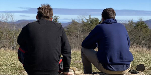Two people sit side by side on a grassy hillside, facing away and looking out at distant trees, hills, and a blue sky. One wears a black sweatshirt, the other a navy hoodie and tan pants.