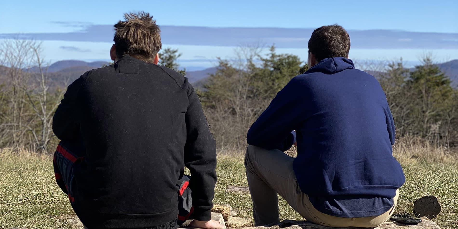 Two people sit side by side on a grassy hillside, facing away and looking out at distant trees, hills, and a blue sky. One wears a black sweatshirt, the other a navy hoodie and tan pants.