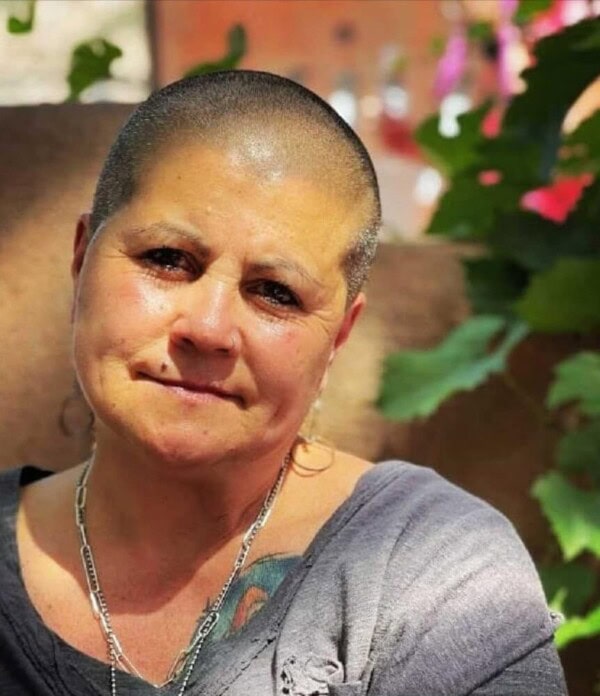 A woman with a shaved head and a light gray top sits outdoors, smiling gently. She wears a silver necklace and has a visible tattoo on her chest. Green leaves and colorful blurred background add to the natural setting.